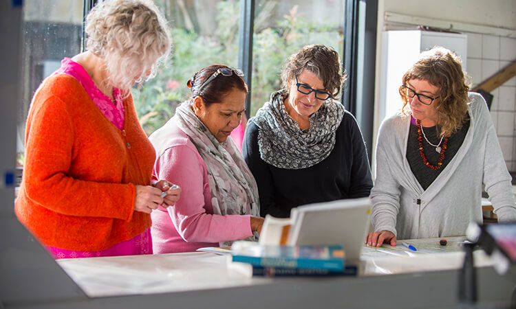 Four women engaged in discussion while looking at documents on a table.