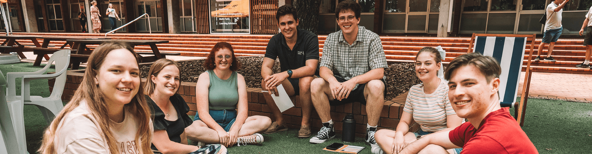 A group of seven people sitting and smiling outside on a lawn near a building.