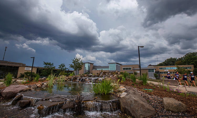 A landscaped area with waterfalls and rocks under a dramatic cloudy sky, with people gathered in the background near a building.