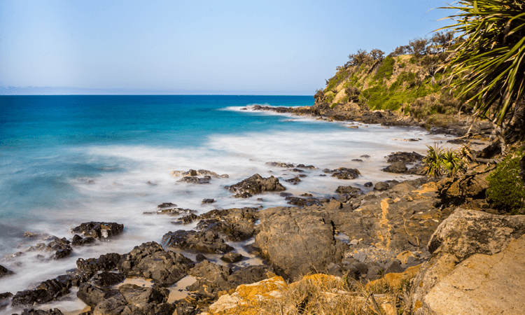 Rocky shoreline with turbulent waves and lush vegetation under a clear sky.