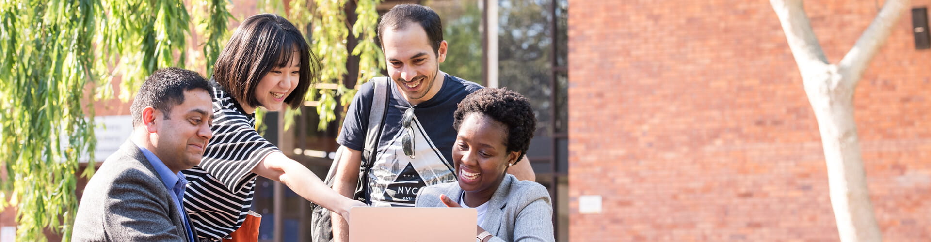 A diverse group of students engaged in a discussion while looking at a laptop outdoors.