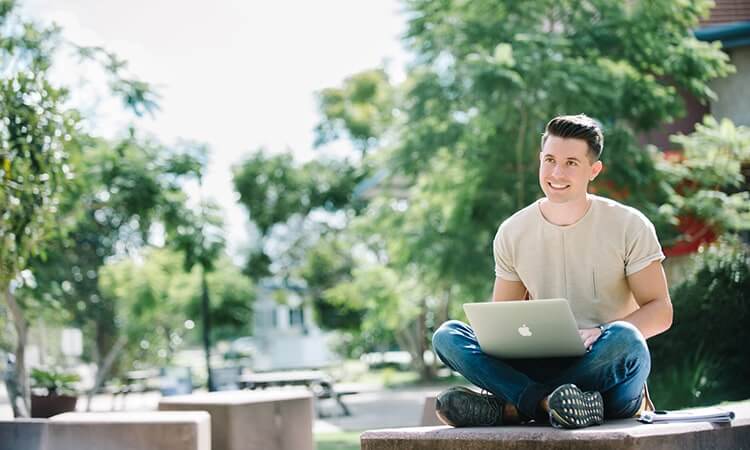 Young man using a laptop while sitting outdoors on a sunny day.