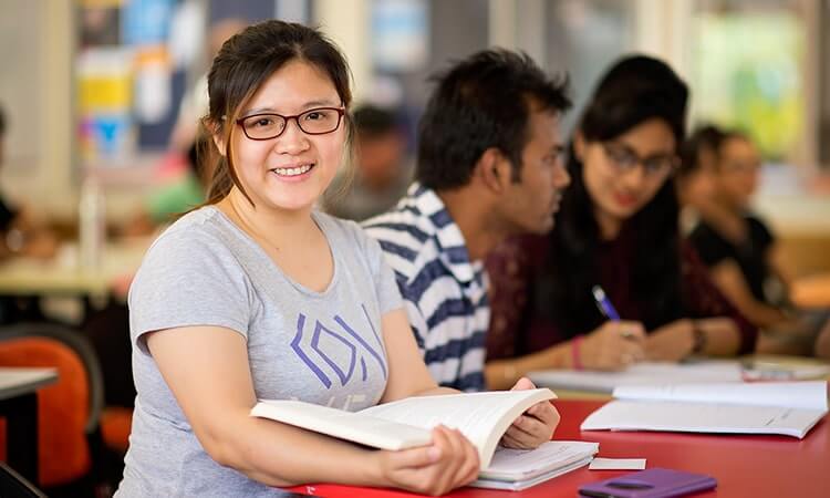 A student smiling at the camera while studying with peers in the background.