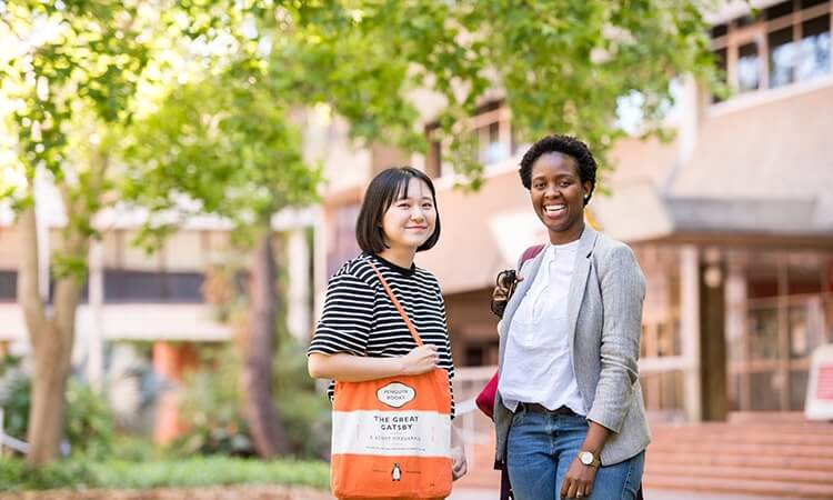 Two smiling women standing outside on a campus with trees and a building in the background.