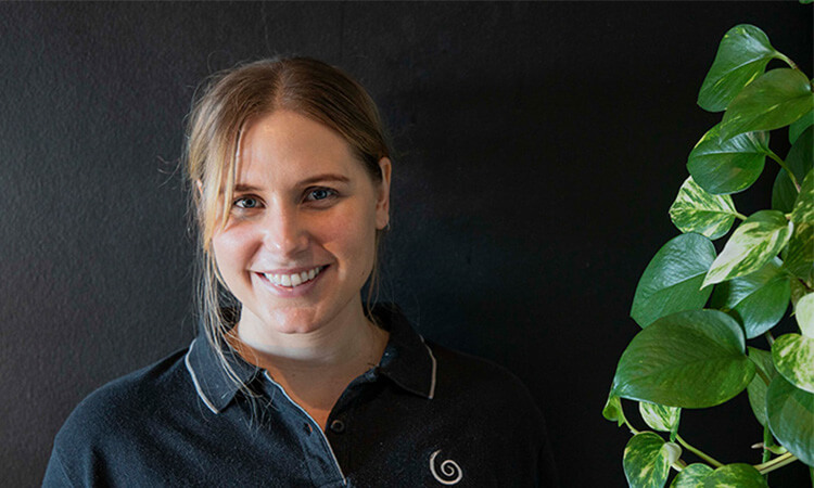 Woman with a smile standing next to a green plant against a black wall.