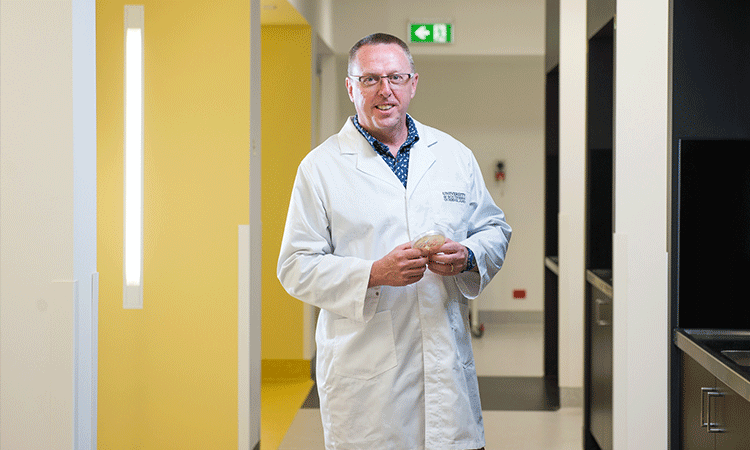 A man in a lab coat standing in a corridor, holding an object in his hands.