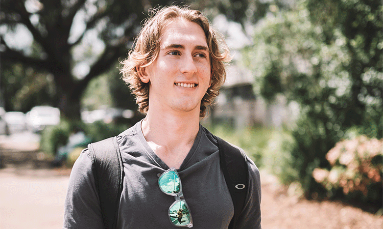 A smiling young man with shoulder-length hair wearing a black t-shirt and sunglasses hanging from his neckline outdoors.