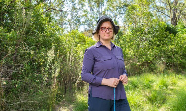 A woman stands in the green outdoors wearing a purple shirt and a hat. She is looking directly at the camera.