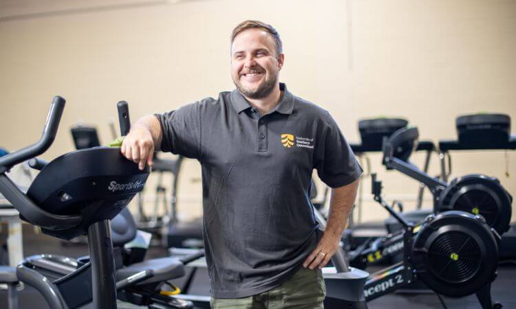 A man, Rhys Brady, stands next to a treadmill in a University of Southern Queensland shirt and exercise shorts. He is smiling.