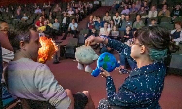 A presenter demonstrates a science concept using plush toys to an engaged audience in a planetarium or lecture hall.