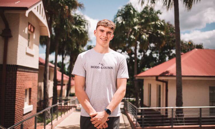 Young man smiling on a sunny day, standing on a bridge with buildings in the background.