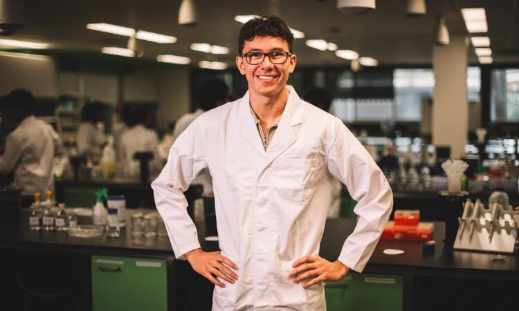 A smiling person in a lab coat standing in front of a laboratory workspace with equipment in the background.