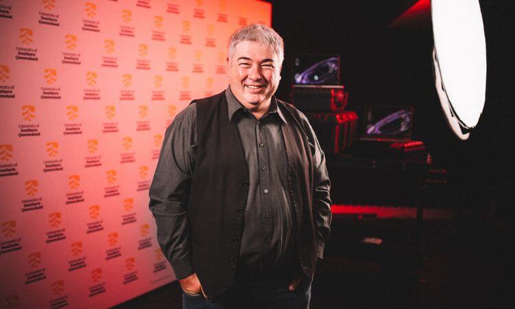 A smiling man in a black shirt and jacket stands in front of a backdrop with logos, with film lighting equipment visible in the background.