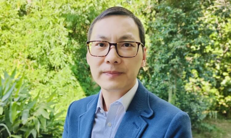 A man wearing glasses, a blue blazer, and striped shirt, standing outdoors with greenery in the background.