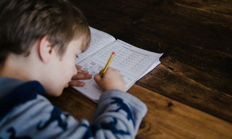 Young boy writing in a workbook with a pencil, sitting at a wooden table, focusing intently on his work.
