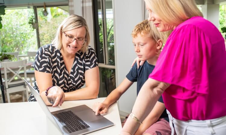 A woman and a young boy seated at a table look at a laptop screen, while another woman standing next to them points at the screen.