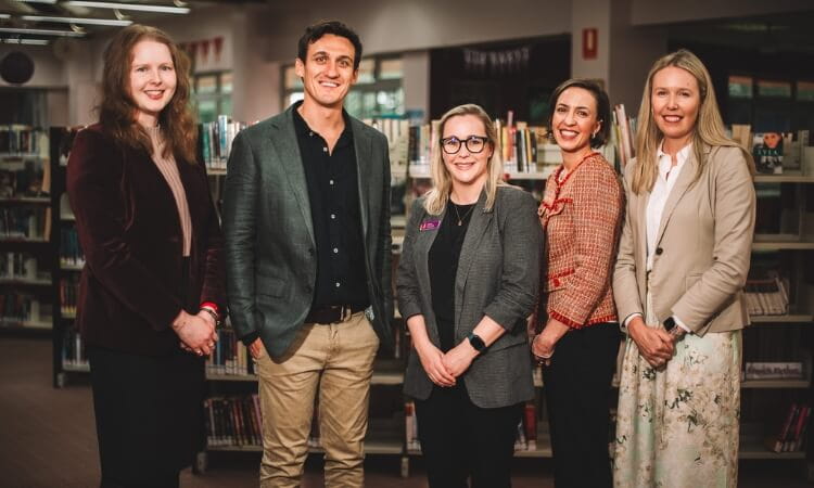 Five professionals smiling in a library, with rows of books in the background. two women on the left, a man in the center, and two women on the right.
