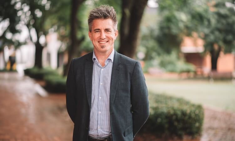 A man in business attire smiling outdoors on a tree-lined path.