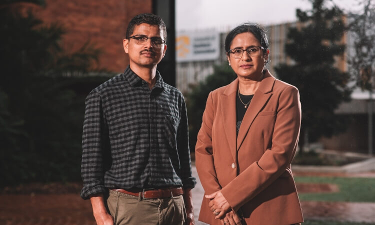 Two professionals standing outdoors, a man in a plaid shirt and glasses, and a woman in a tan blazer, both looking at the camera.