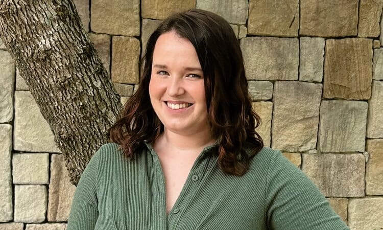 A smiling woman with shoulder-length brown hair, wearing a green blouse, standing in front of a stone wall and a tree.