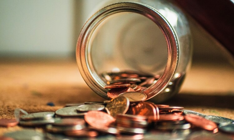 A glass jar tipped over with a pile of scattered coins on a wooden surface.