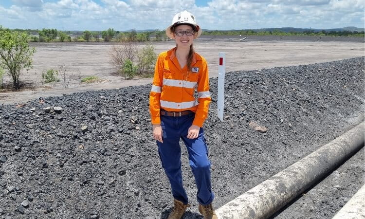 A person wearing a hard hat, safety glasses, and high-visibility shirt standing on a rocky terrain with an open field in the background.