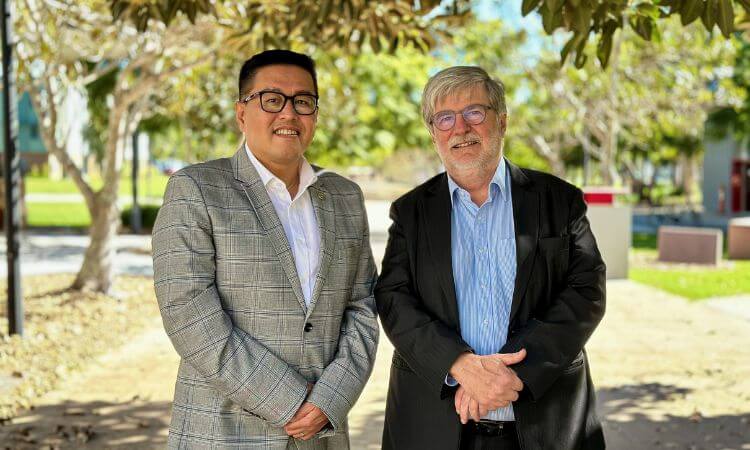 Two men are standing outdoors under a tree. One is wearing a checked blazer and glasses, while the other is dressed in a dark suit and has gray hair and beard. They are both smiling at the camera.