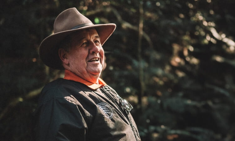 A man wearing a wide-brimmed hat and dark jacket stands outdoors, smiling, with trees and foliage in the background.