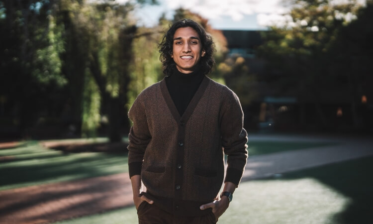 Man poses outdoors wearing a brown cardigan over a black turtleneck. 