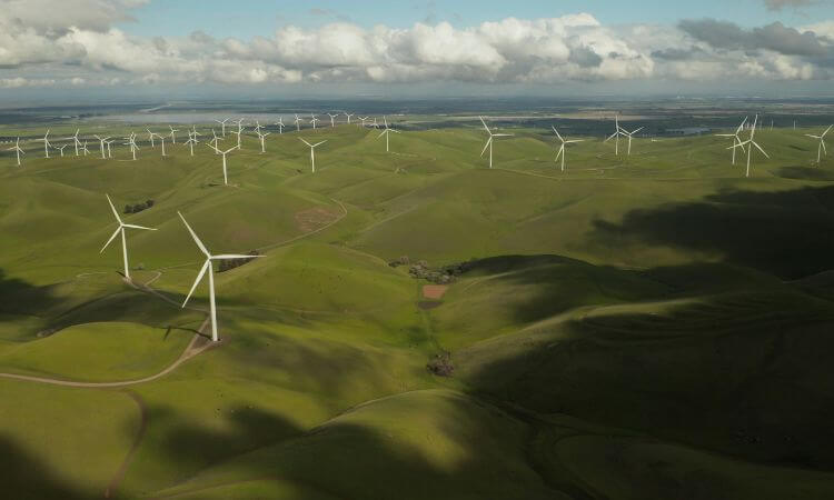 Expansive view of a wind farm with numerous turbines on rolling green hills under a partly cloudy sky.