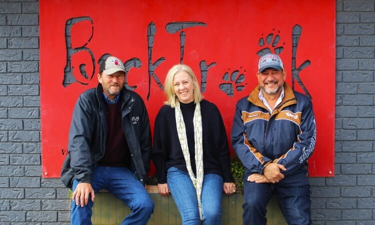 Three people wearing jackets and hats are smiling while sitting in front of a red sign.