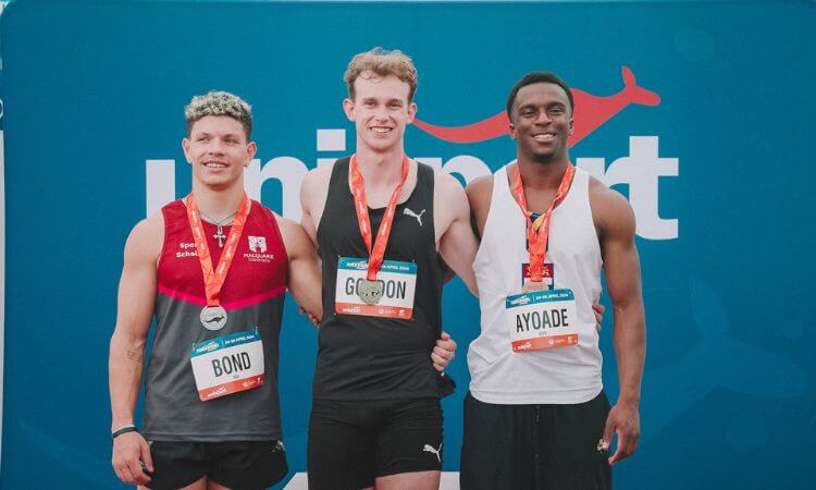 Three male athletes wearing medals stand smiling on a podium with a banner reading "unisport" in the background.