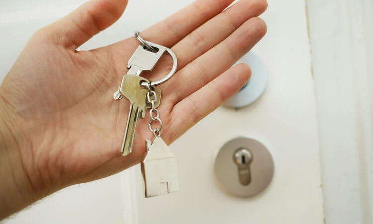 A hand holds a keyring with two keys and a house-shaped keychain, with a door lock in the background.