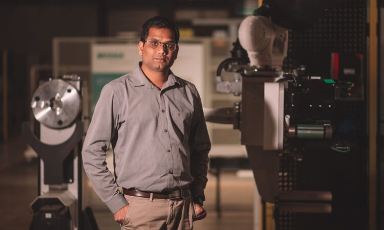 A man in a gray shirt and glasses stands in an industrial setting with machinery and equipment in the background.