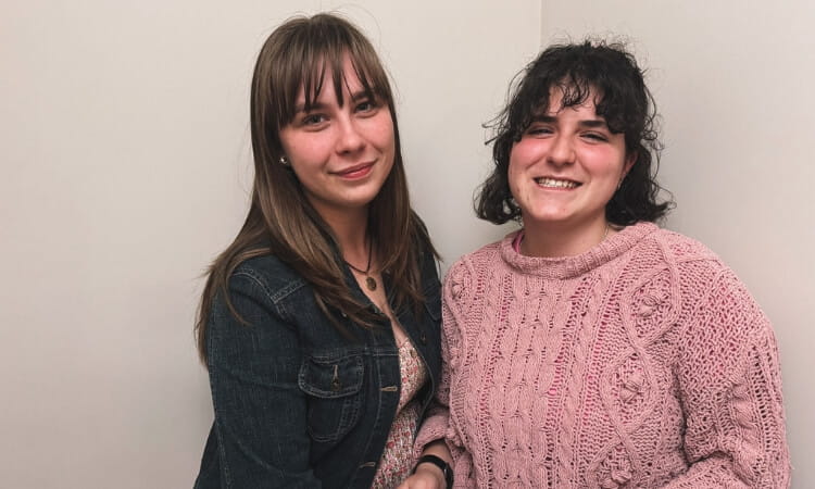 Two women standing close together, one wearing a denim jacket and the other in a pink knitted sweater, both smiling at the camera.