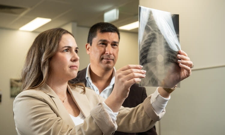 Two individuals in a medical setting examining an X-ray image held up to the light, focusing on the details shown on the film.