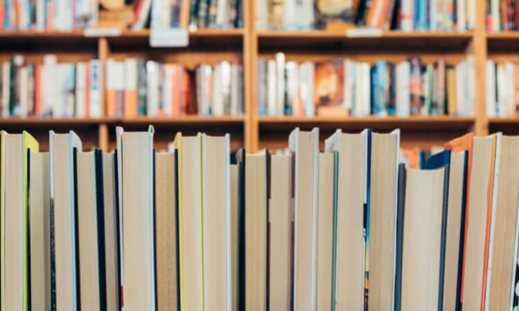 A row of books on a shelf, spines turned away, with a blurred background showing more bookshelves filled with various books.