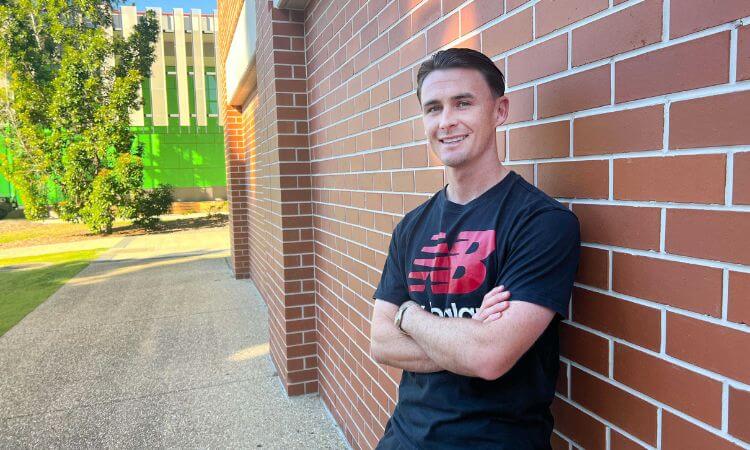 A person in a black New Balance shirt stands smiling with arms crossed against a brick wall. A green and white building is in the background.