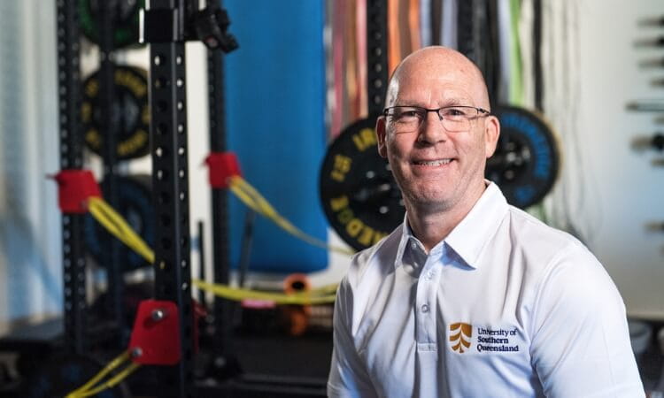 A man wearing glasses and a white polo shirt smiles. He is seated in front of gym equipment.