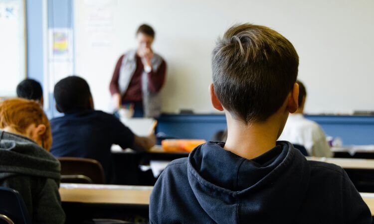 Students sitting at desks in a classroom, with a teacher standing at the front near a whiteboard.