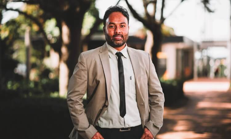 A man wearing a beige suit and black tie is standing outdoors with trees and buildings blurred in the background.