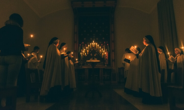 A group of people holding candles in a dimly lit room, standing and reading from books, facing an altar adorned with multiple lit candles.
