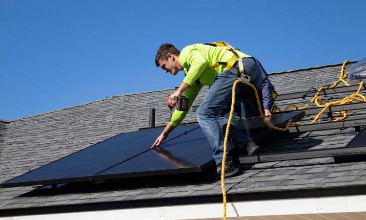 A worker in a bright green shirt installs solar panels on a residential roof under a clear blue sky.