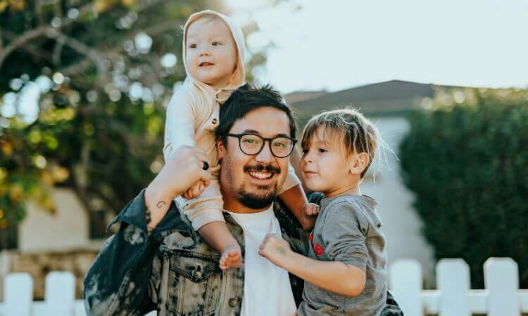 A man smiling while holding a baby and standing next to a child outdoors. A white picket fence and trees are in the background.