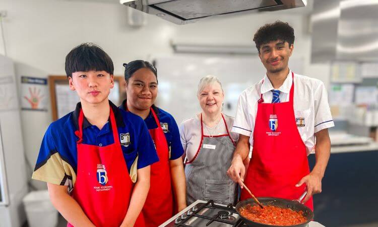 Four people in red aprons stand in a kitchen, three are teenagers and one is an older adult. They are gathered around a stove with a pan of food.