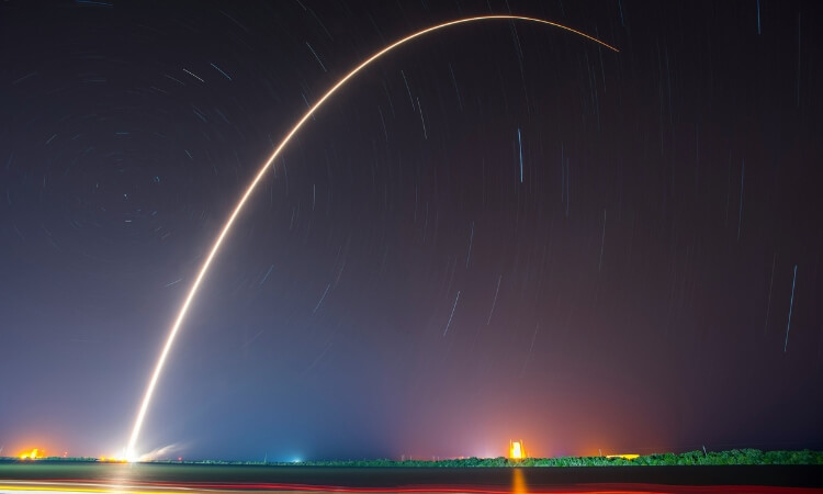 A long-exposure photo shows the arc of a rocket launch at night, with a bright trail against a dark sky and distant lights on the horizon.