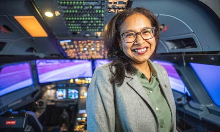 A person in a gray blazer stands smiling inside an aircraft cockpit with glowing control panels and screens in the background.