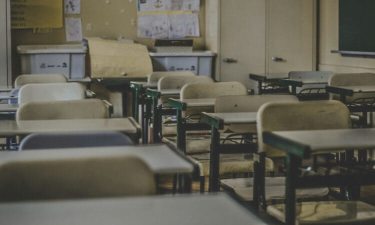 An empty classroom with rows of desks and chairs, light-colored walls adorned with papers, and a chalkboard in the background.