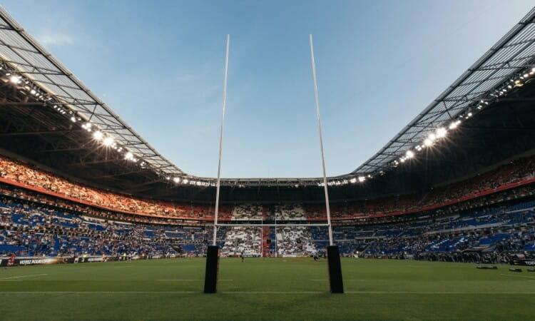 Wide view of a stadium with rugby goalposts in the foreground, surrounded by spectators in the stands under a clear sky.