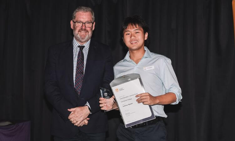 Two people standing side by side. One holds an award and a certificate. Both are smiling, dressed in formal attire, and standing against a dark curtain backdrop.